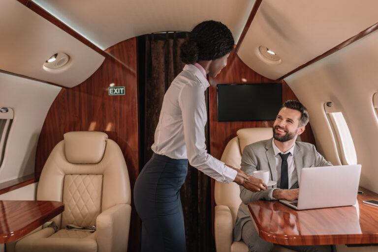 young african american stewardess giving cup of coffee to smiling businessman sitting at laptop in